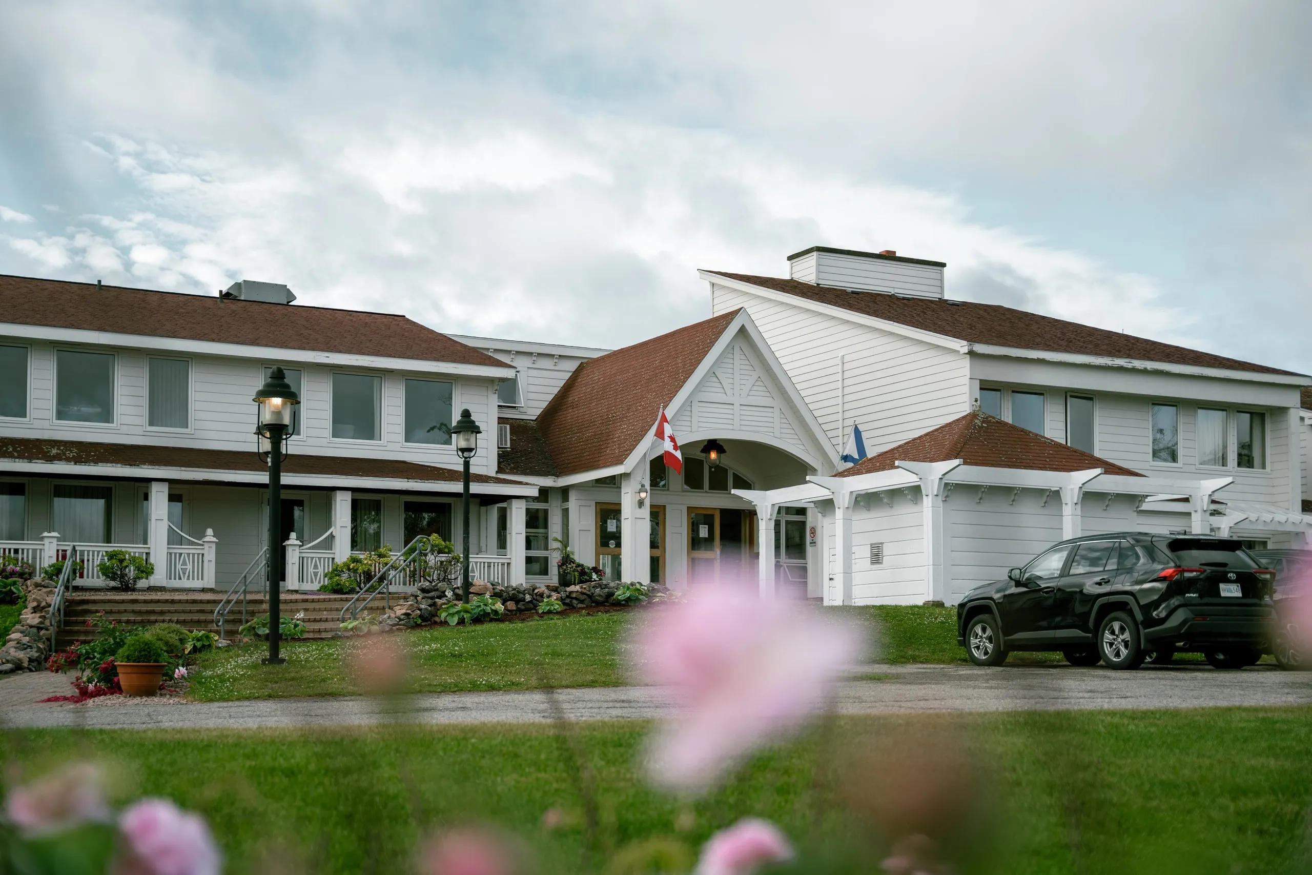 The Keltic Resort’s white lodge entrance, with the Canadian flag flying, a black SUV under the covered drive, and pink flowers in the foreground.