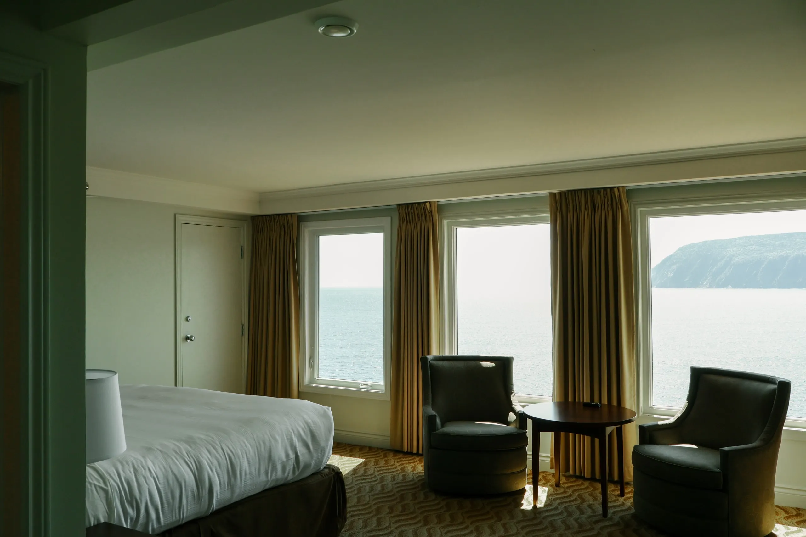 A guest room with two armchairs facing tall windows onto the ocean and a forested headland.