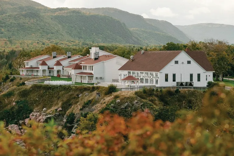 The Keltic Resort lodges with red roofs on a rocky cliff, autumn foliage in front and forested mountains behind.