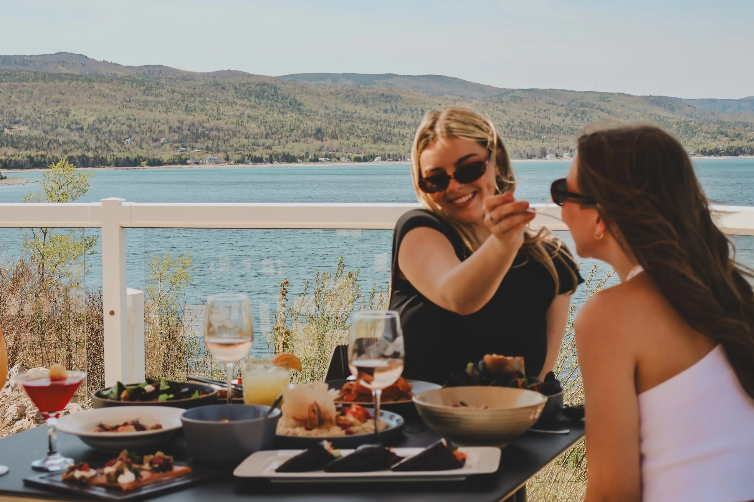 Two people toasting with wine at an outdoor table overlooking the water and hills.