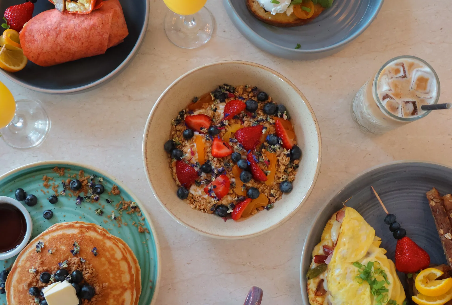 A breakfast table from above, with yogurt, berries, and granola, pancakes, an omelette, and iced coffee.