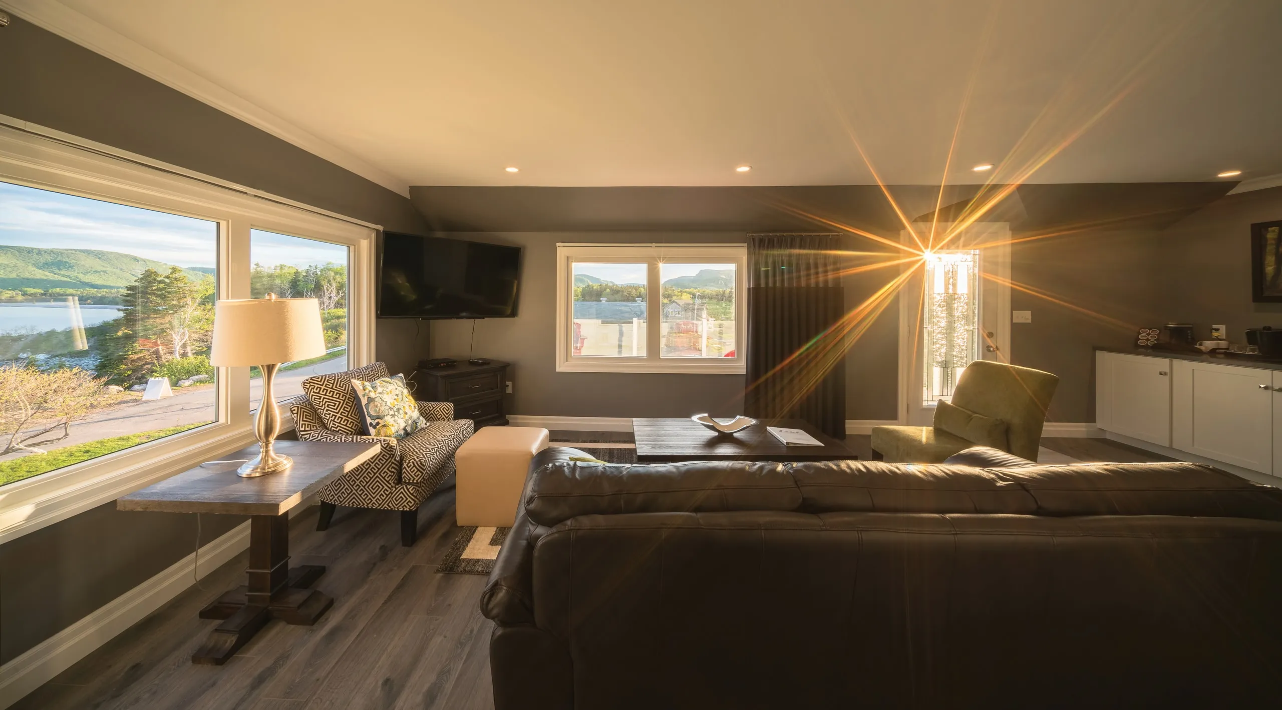 A living room at sunset with a dark sofa facing windows over water and forested hills.