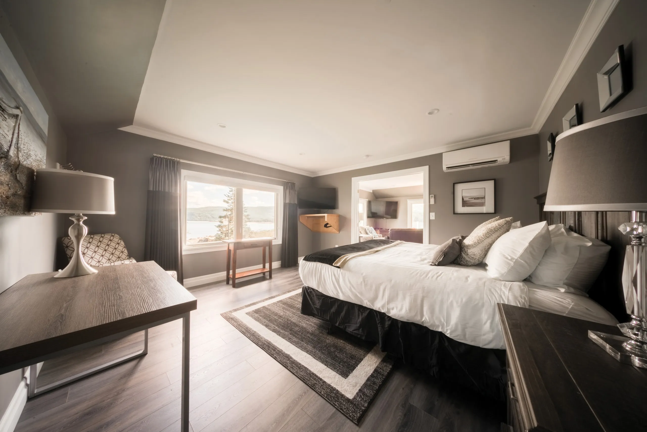 A bedroom with white linens, a desk lamp in the foreground, and a window looking onto the landscape.