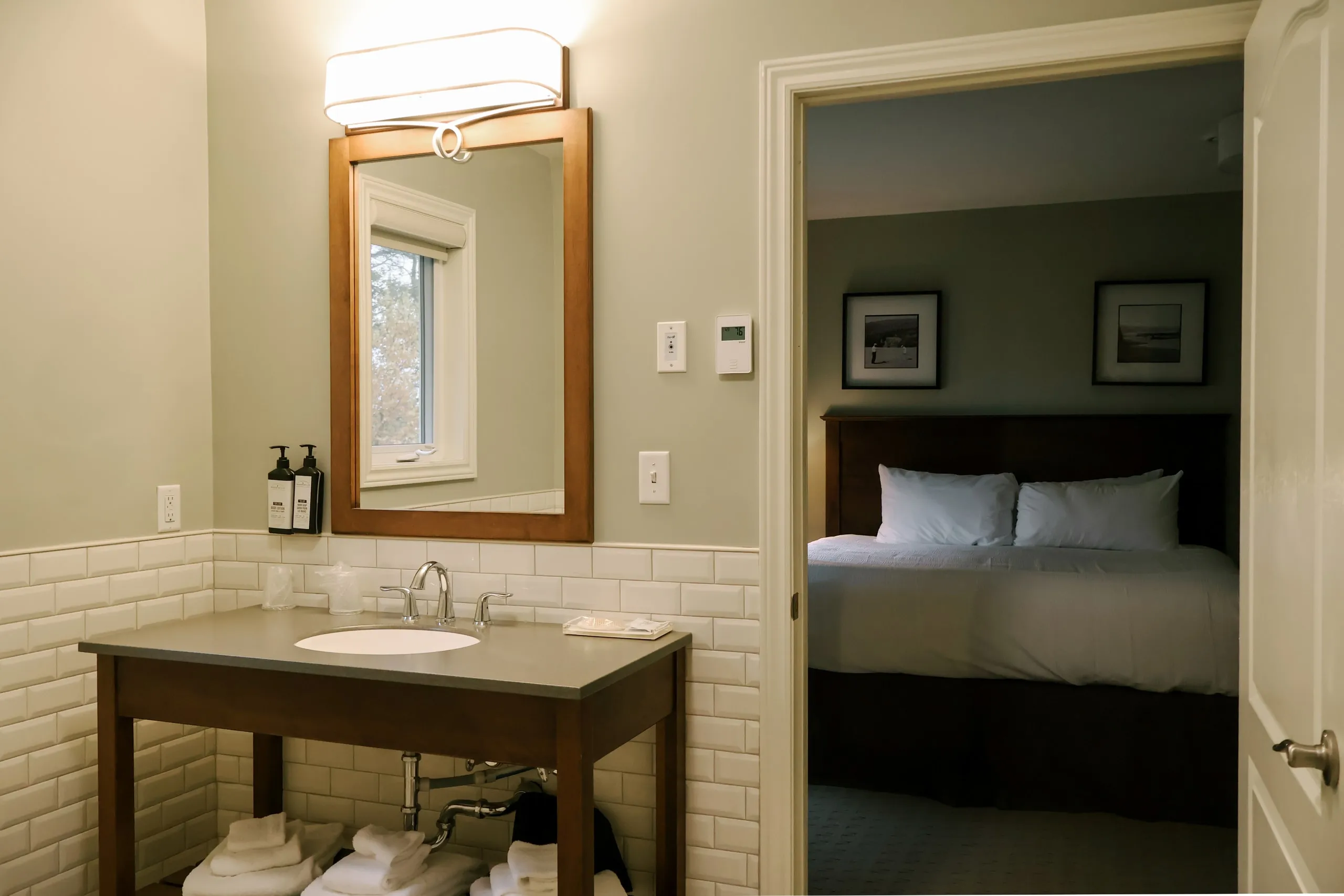 An ensuite bathroom vanity with subway tile and a wood-framed mirror, looking through to the bedroom.