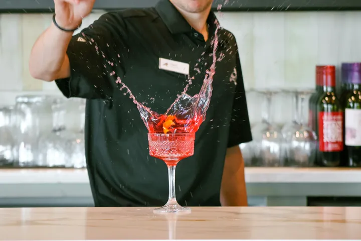 A bartender in a black polo sending a red cocktail splashing from a stemmed glass on a wooden bar.