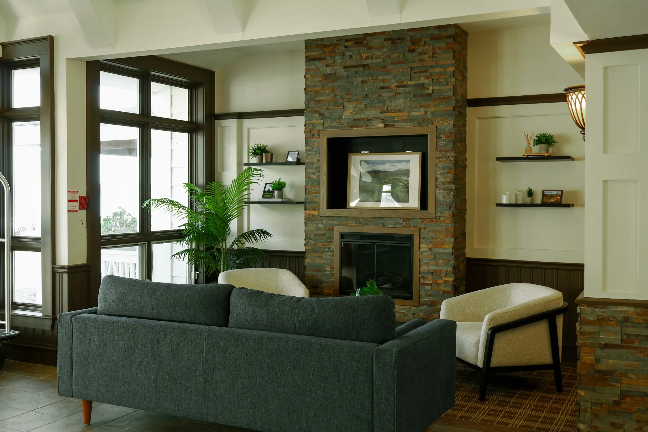 A lounge with a stone fireplace, a grey sofa, and chairs beside tall windows.