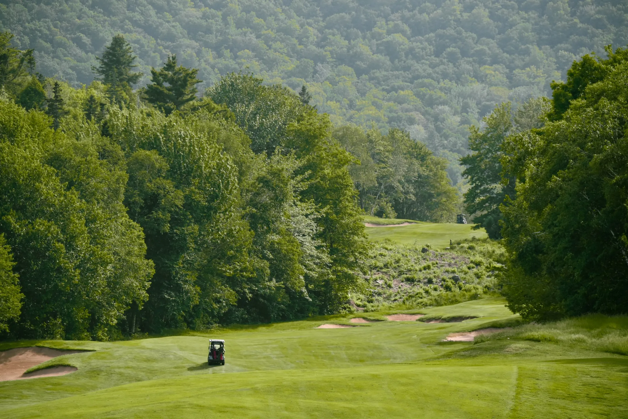 A golf cart on a fairway lined with bunkers, with forested Highlands rising behind the green.