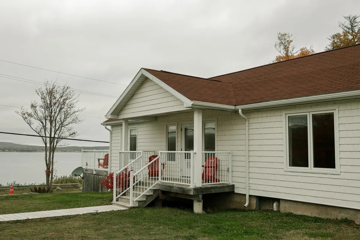 A white clapboard suite with a brown roof and a deck of red chairs beside the water.