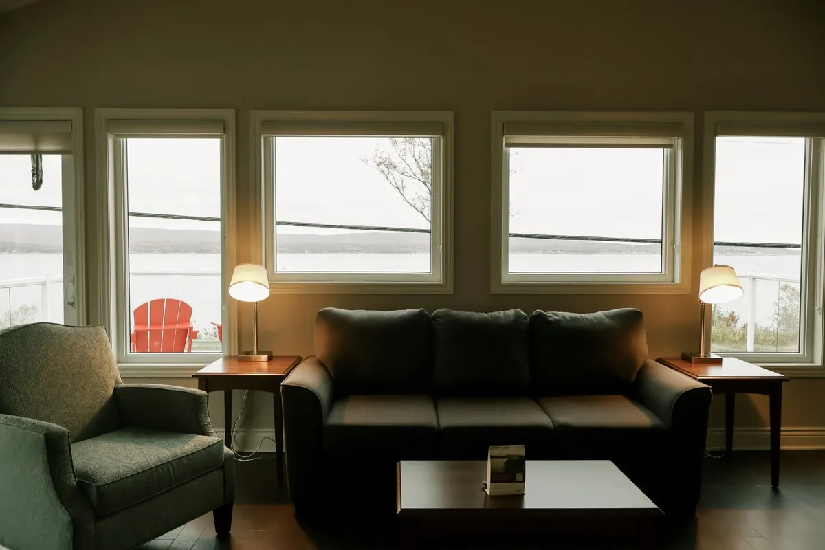 A living room with a dark sofa, a green armchair, and windows looking onto the water and a red chair.