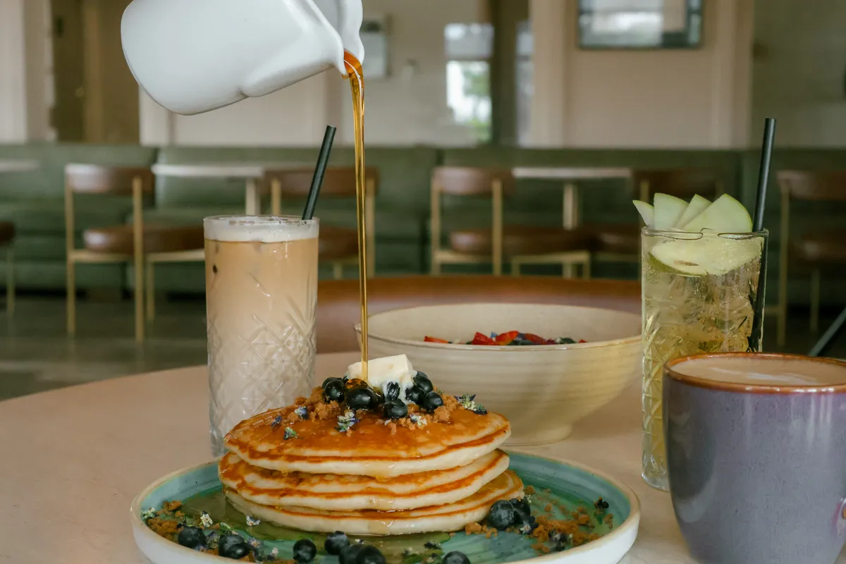Maple syrup poured over blueberry pancakes beside an iced coffee, fruit bowl, and apple drink.