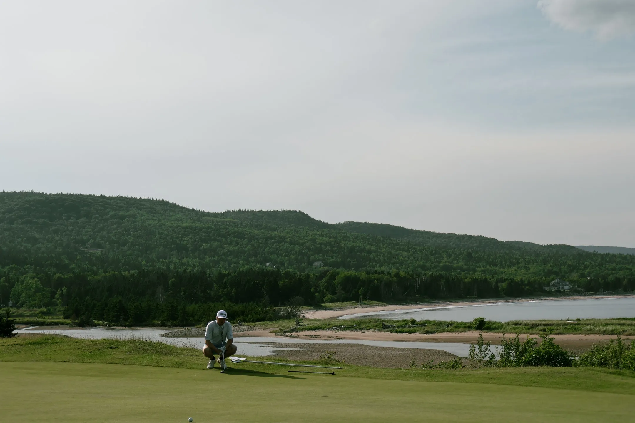 A golfer lining up a putt on a green above a tidal river, beach, and forested hills.