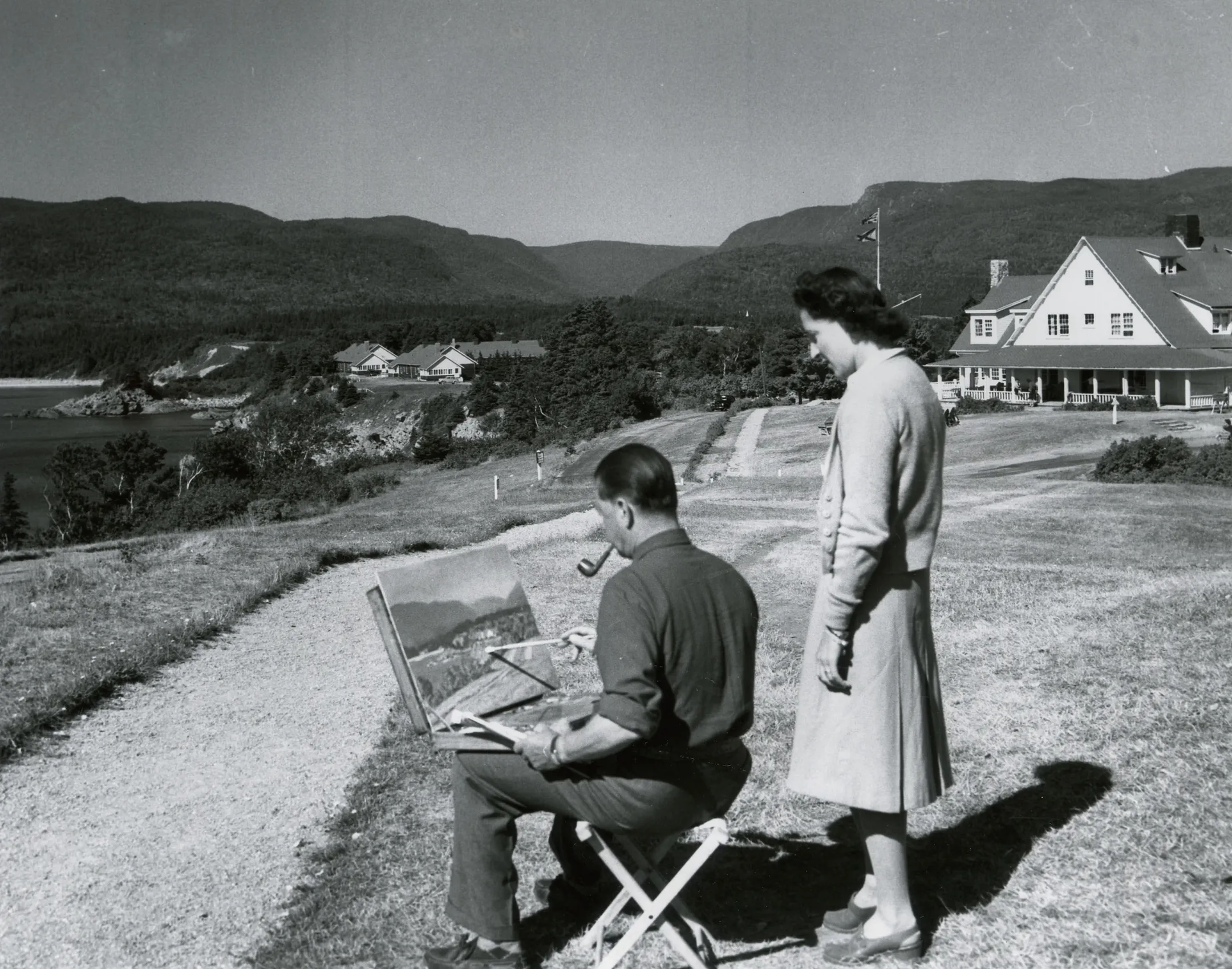 Archival photograph of a man painting outdoors at an easel with a woman watching beside him.