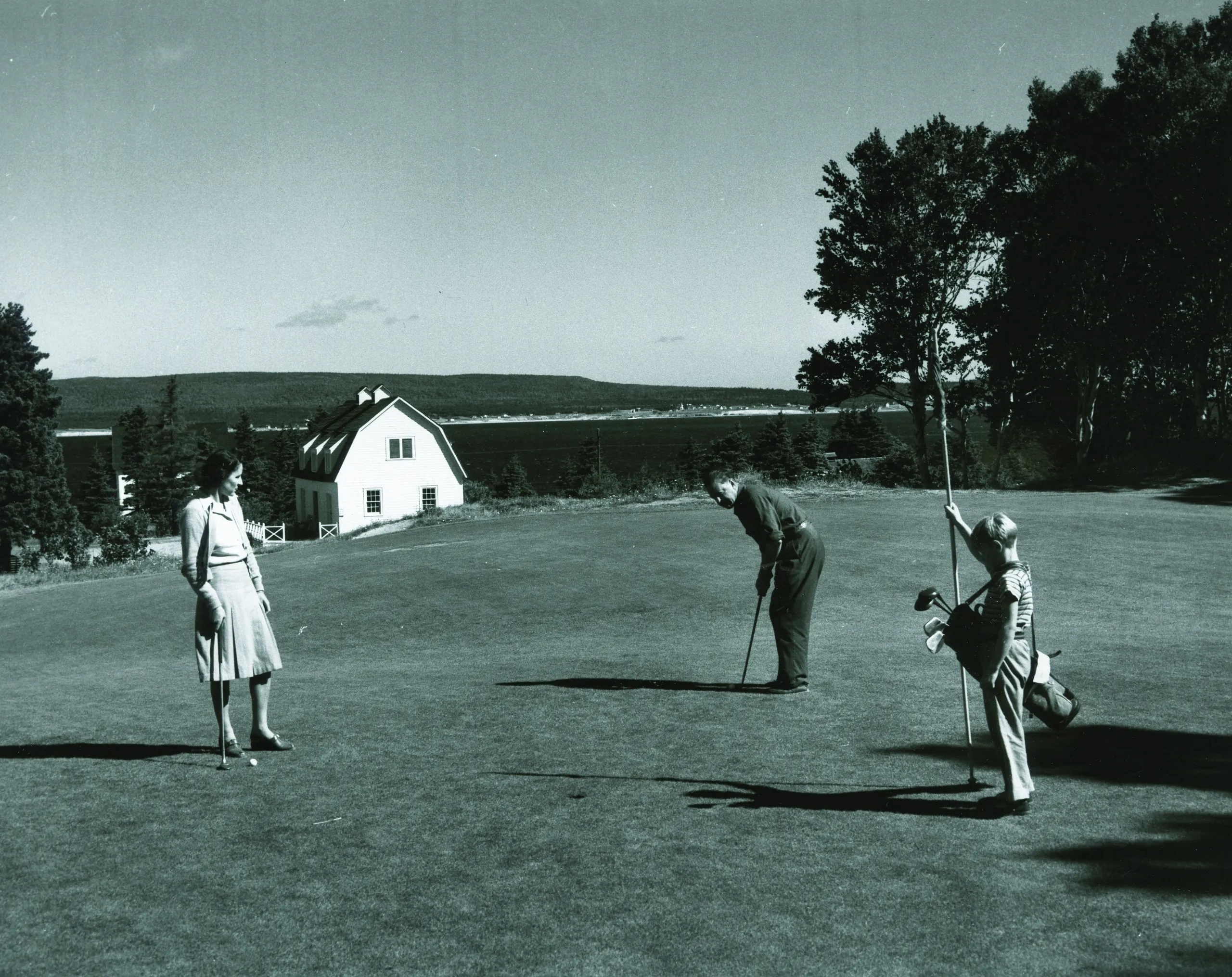 Archival photograph of three people golfing on the Highlands Links course with the lodge and Atlantic coastline in the background.