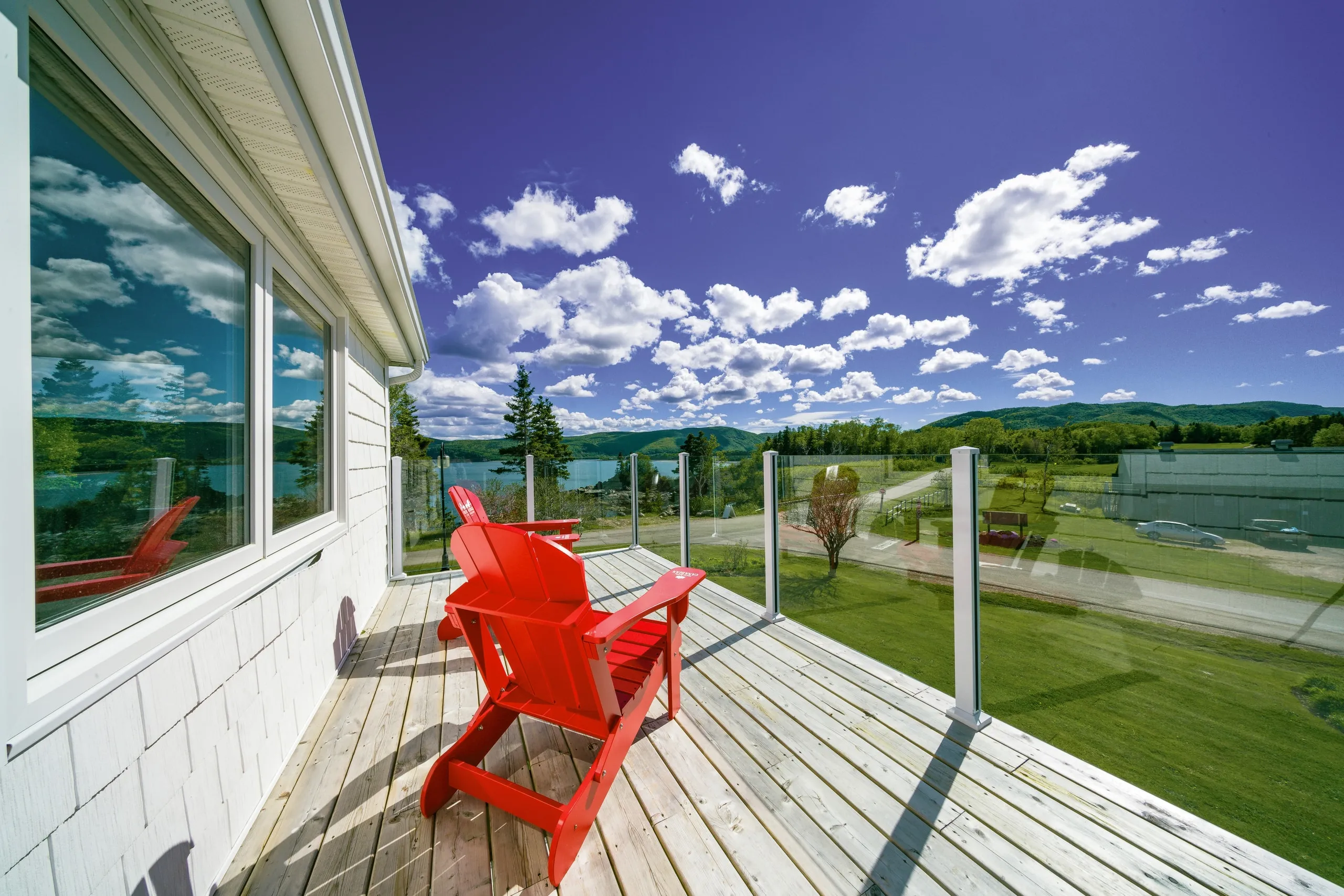 Two red Adirondack chairs on a wooden deck with a glass railing overlooking water and green hills.