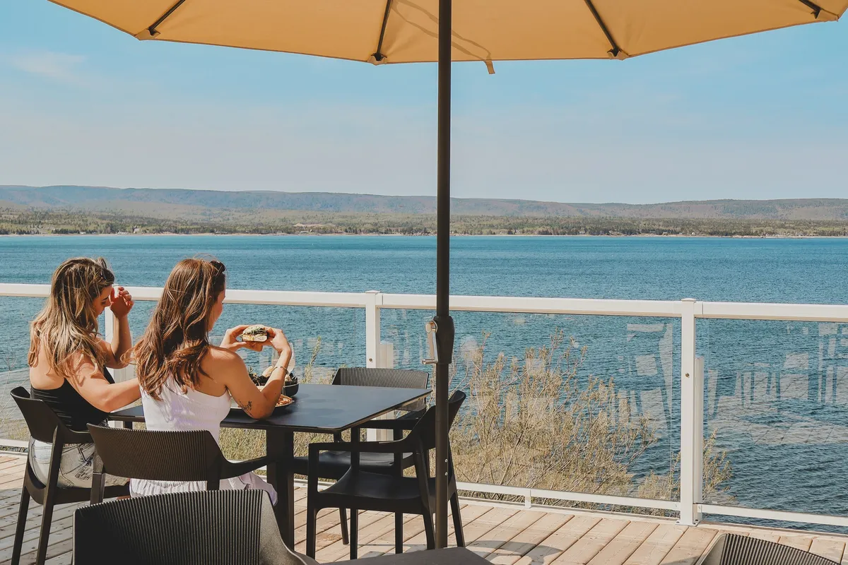Two guests seated at an oceanfront patio table under a beige umbrella, looking out over the water.