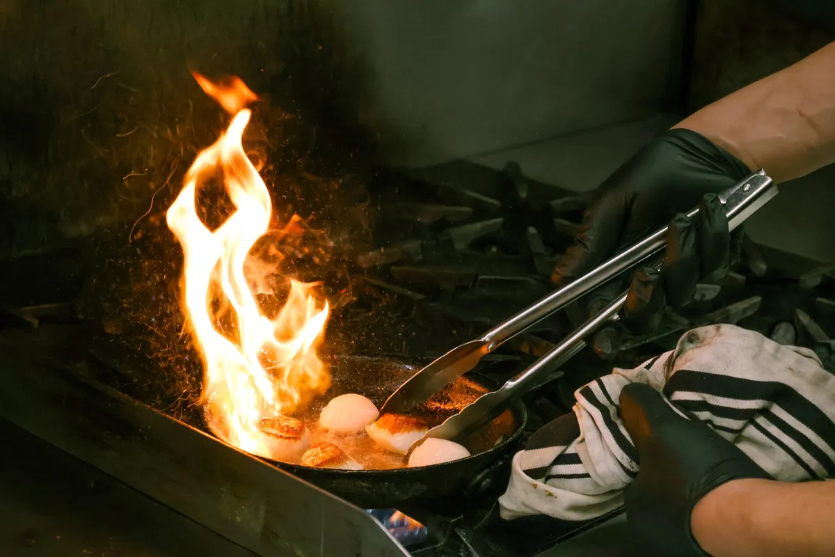 A chef flambéing scallops in a pan with tongs.