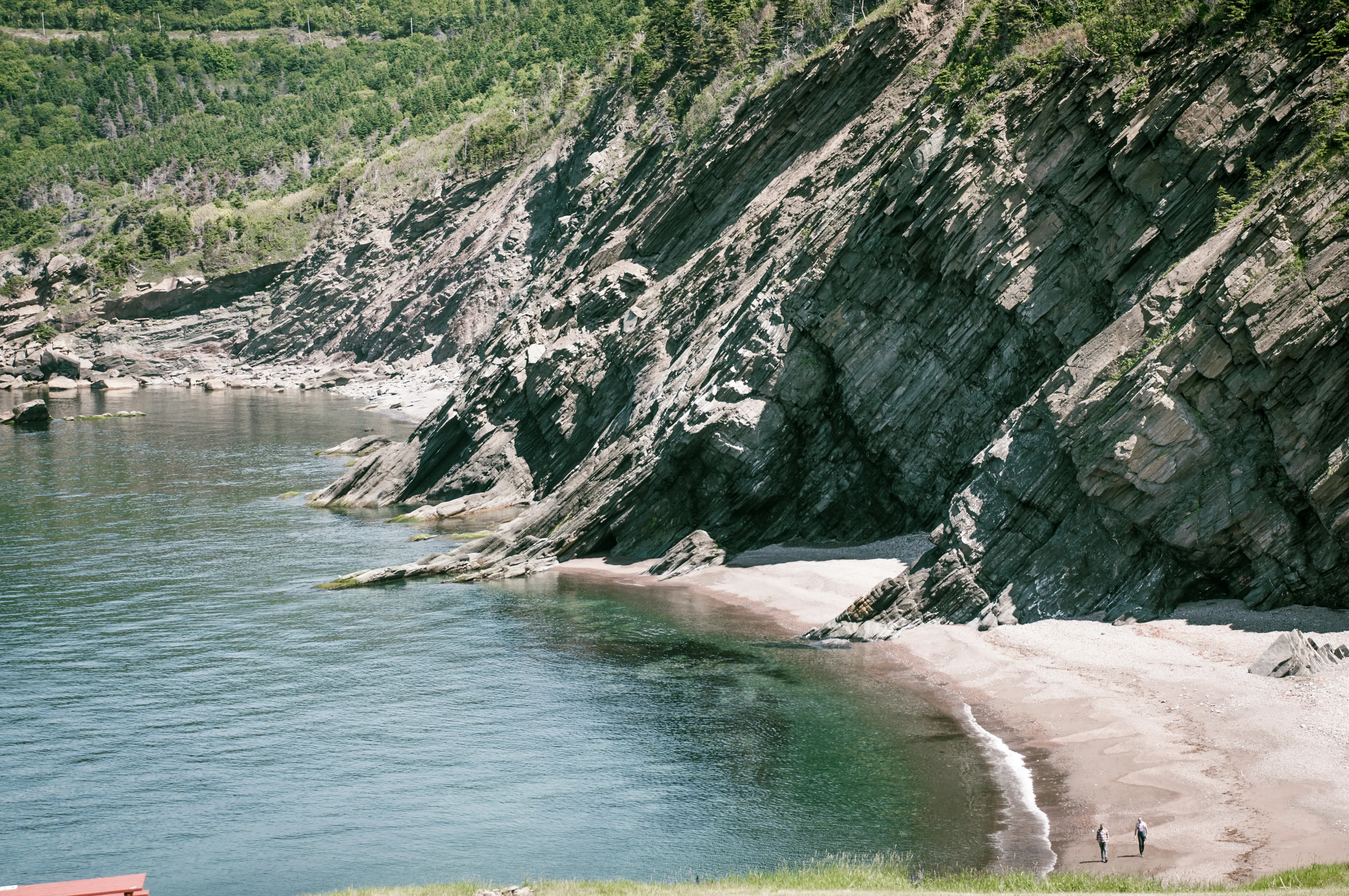 Two people walking a sandy cove beneath steep layered cliffs along the coast.