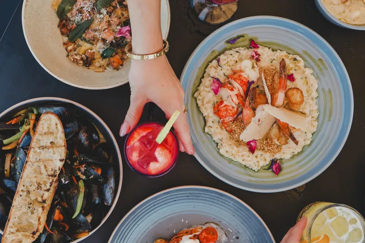 An overhead view of a table with lobster risotto, mussels, grilled bread, and a hand reaching for a red cocktail.