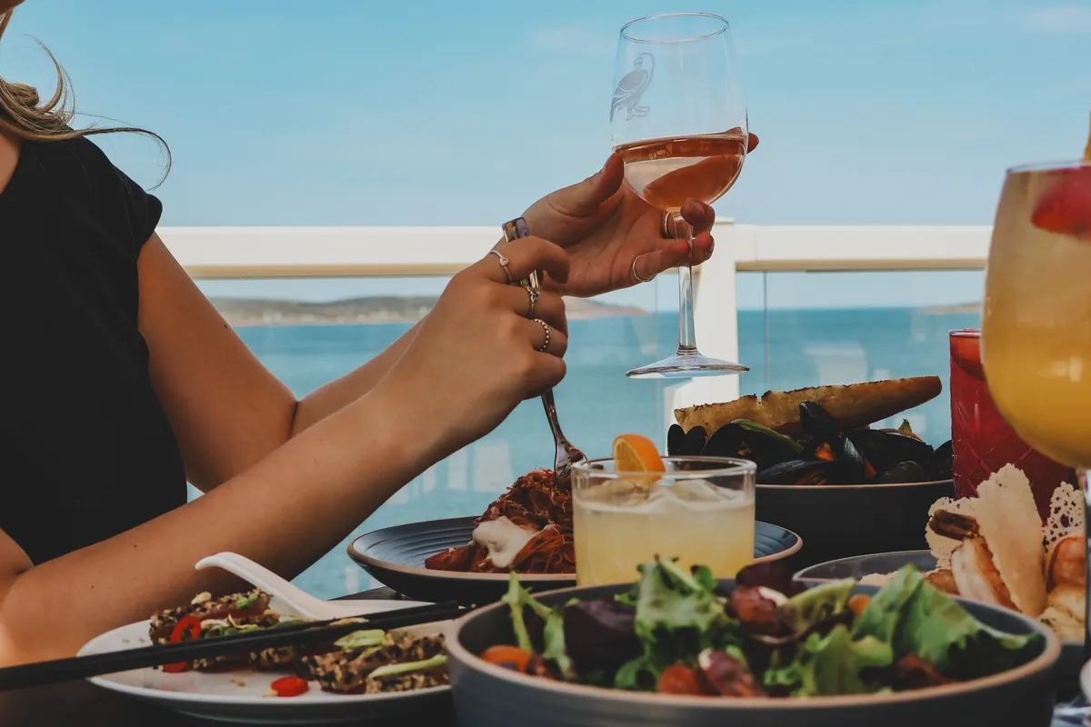 Hands toasting a glass of rosé over plates of seafood and salad on a patio overlooking the water.