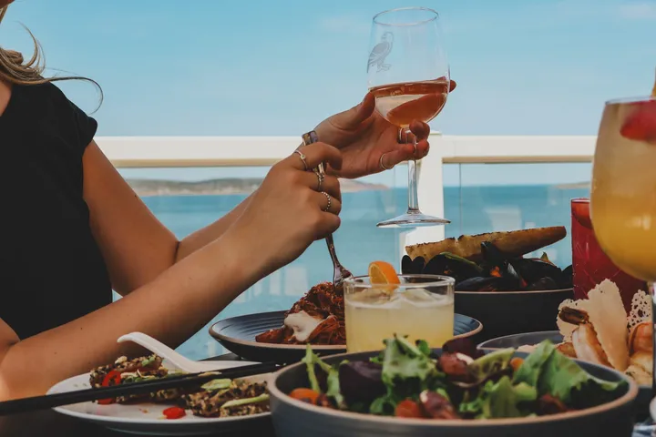 Hands toasting a glass of rosé over plates of seafood and salad on a patio overlooking the water.